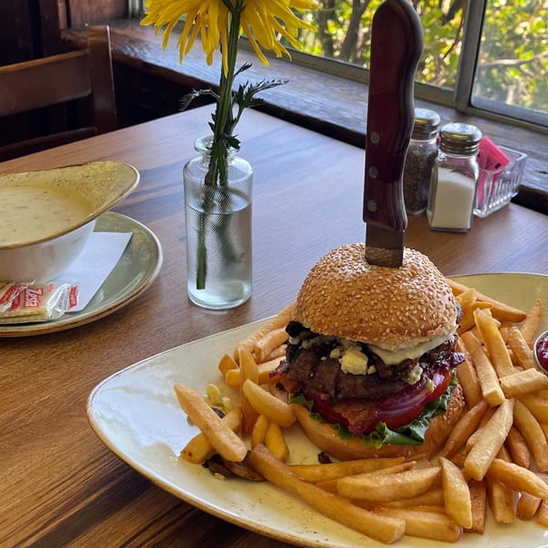 A burger with a knife, fries, and a bowl of soup on a wooden table by a window.