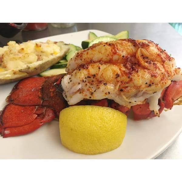 Grilled lobster tail with a baked potato, lemon wedge, and sliced cucumbers on a white plate.