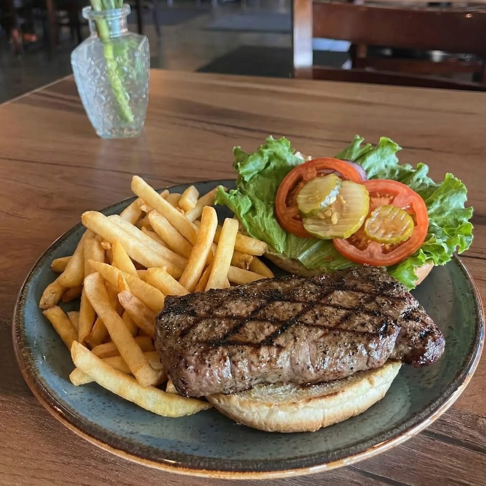 Grilled steak sandwich with lettuce, tomato, pickles, and a side of french fries on a blue plate.