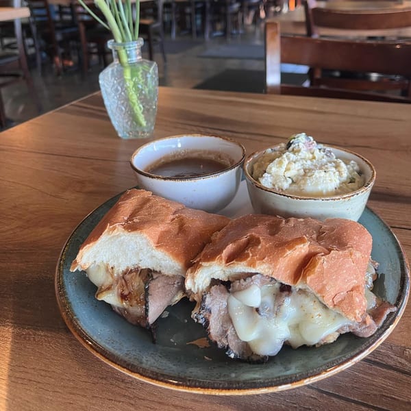A French dip sandwich with au jus and potato salad on a wooden table at The Sardine Can.