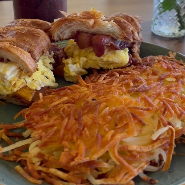 Two croissant breakfast sandwiches with scrambled eggs and bacon, alongside crispy hash browns.