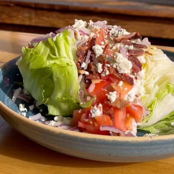 A wedge salad with lettuce, tomatoes, red onions, bacon bits, and feta cheese in a blue bowl.